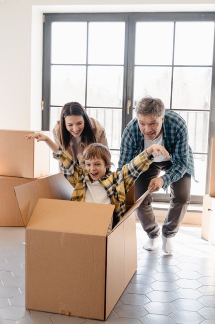 A smiling man and woman are engaged in a home relocation process outside a property, with the man holding a cardboard box with a red 'Fragile' handle with care sticker on it, and the woman holding ano